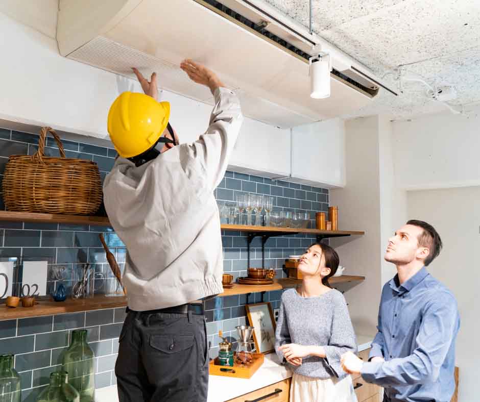A technician in a yellow hard hat inspects a ceiling air conditioner in a modern kitchen